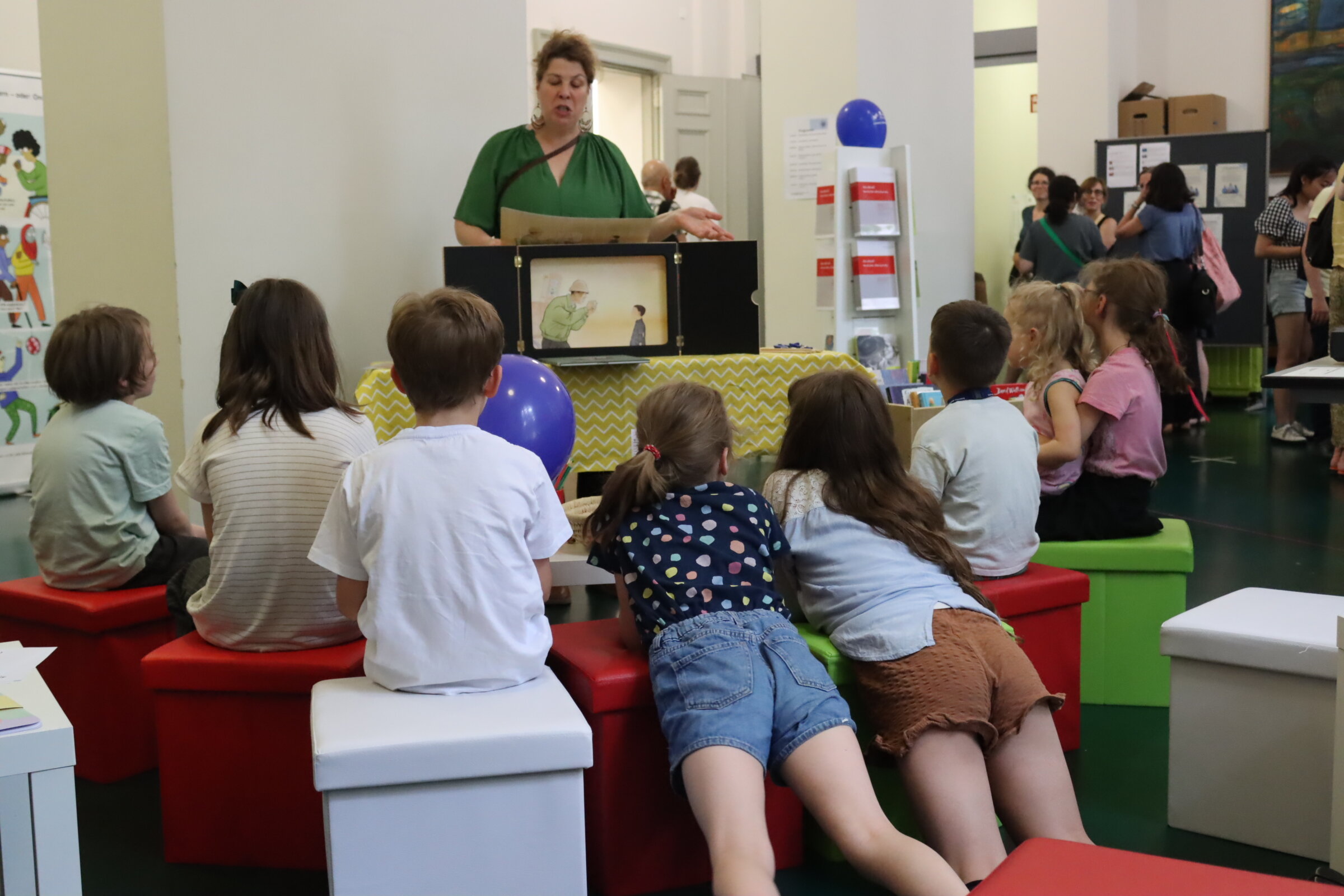 Das Foto zeigt eine Mitarbeiterin der kooperierenden Bibliothek, die mittels eines Kamishibai-Theaters eine Geschichte erzählt. Davor sind acht Kinder zu sehen, die sitzend oder auf Sitzhockern lehnend, gespannt der Geschichte lauschen.