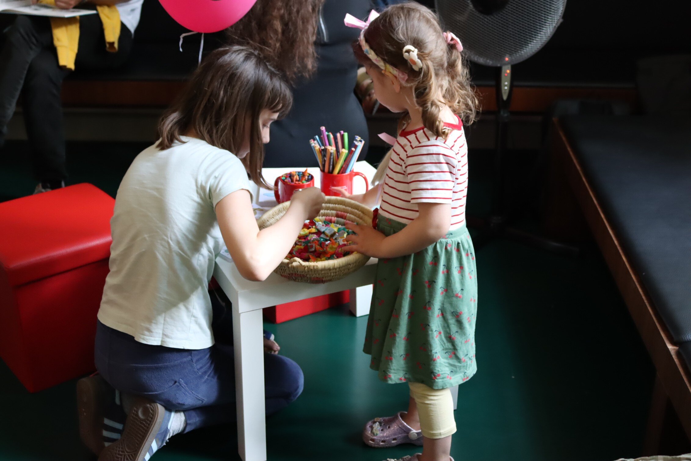 Zwei Kinder stehen bzw. knien an einem niedrigen Tisch, auf dem sich Buntstifte und eine Schale mit Bonbons befinden.