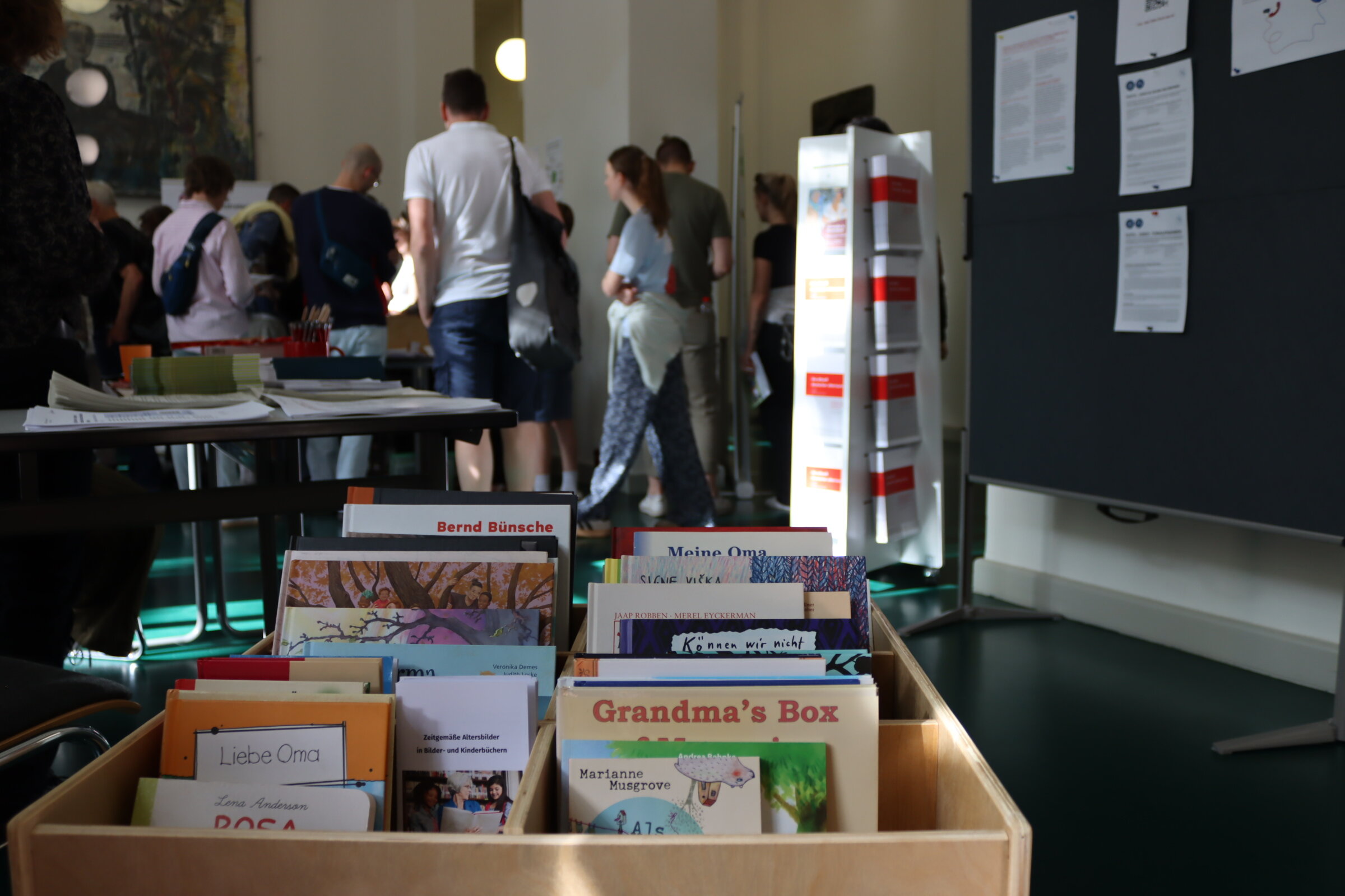 Im Vordergrund steht ein Holzbox mit verschiedenen Bilder- und Kinderbüchern. Weiter hinten steht ein Buchaufsteller mit verschiedenen Ausgaben des DZA Aktuell. Zudem sieht man im Hintergrund Besucher*innen der Veranstaltung.
