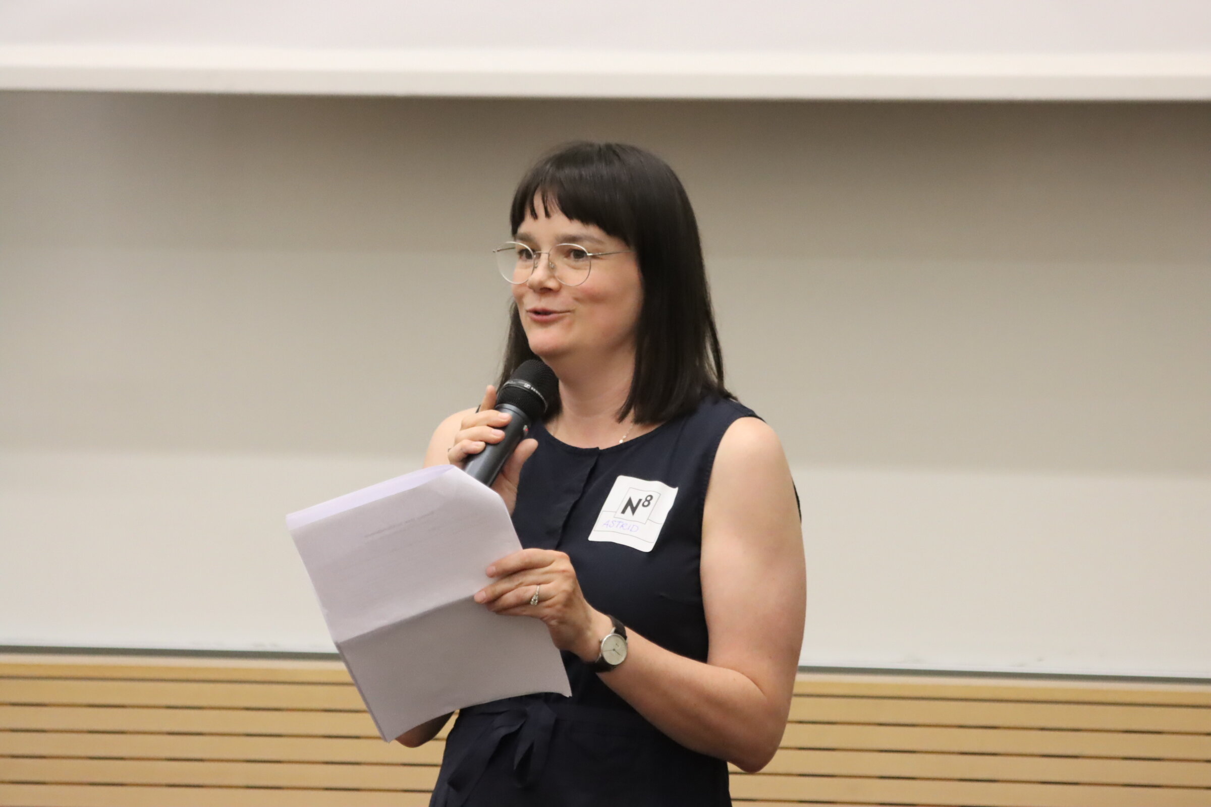 Astrid L&auml;rm steht mit einem Mikrofon und Moderationskarten in den H&auml;nden im H&ouml;rsaal.