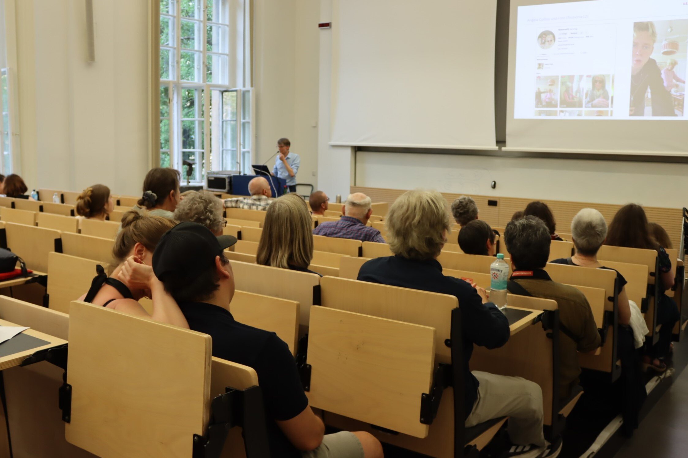 Das Foto zeigt den Hörsaal mit den zahlreichen Besuchern in Rückenansicht. Zu sehen ist auch eine Präsentation, auf der ein Video eines Granfluencers eingebettet ist.