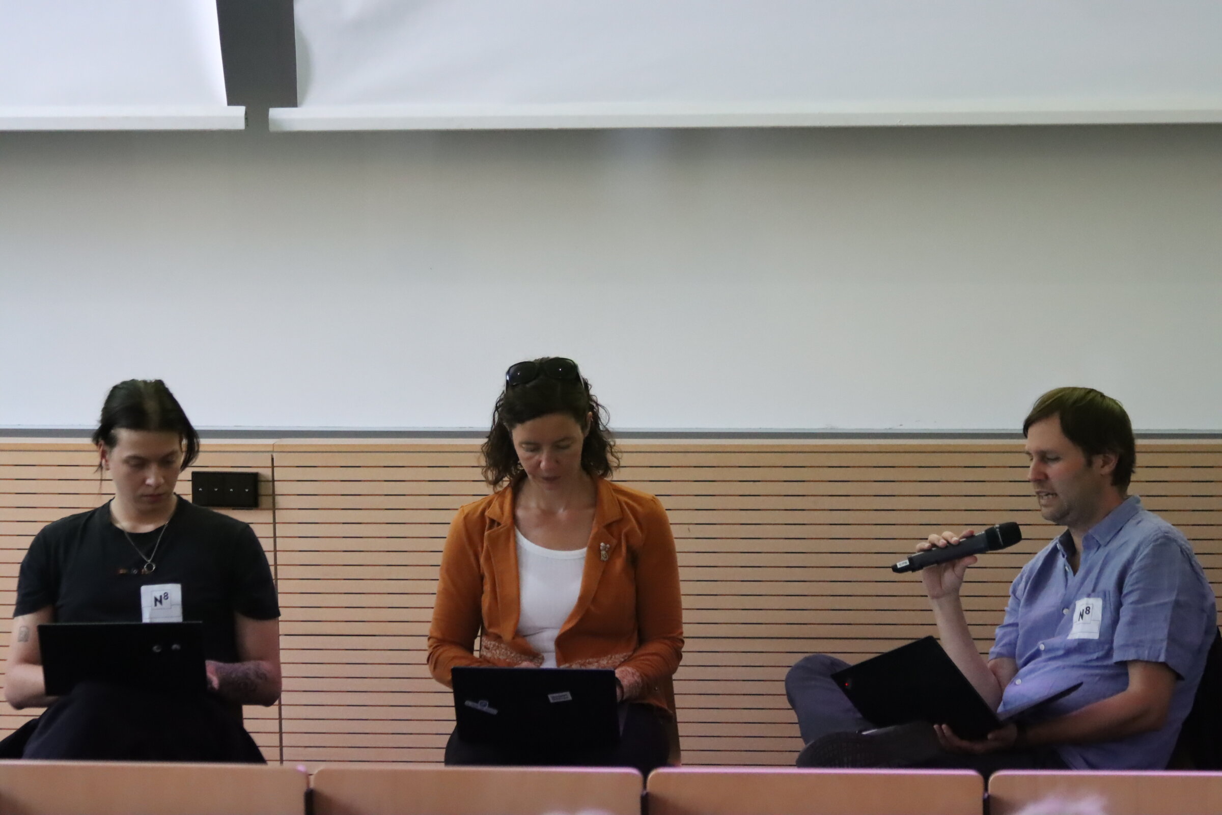 Drei Mitarbeiter*innen des DZA sitzen neben dem Rednerpult im Hörsaal. Sie halten Laptops auf den Knien und rechnen. Ein Kollege hält ein Mikrofon in der Hand und berichtet Ergebnisse.