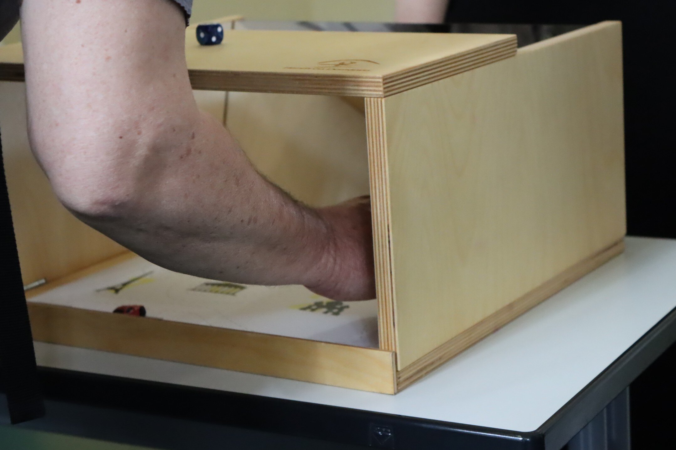 Zu sehen ist eine Hand, die in einer Holzbox-Spiegelkonstruktion steckt. Es geht darum unter erschwerten Bedingungen etwas zu Malen. 