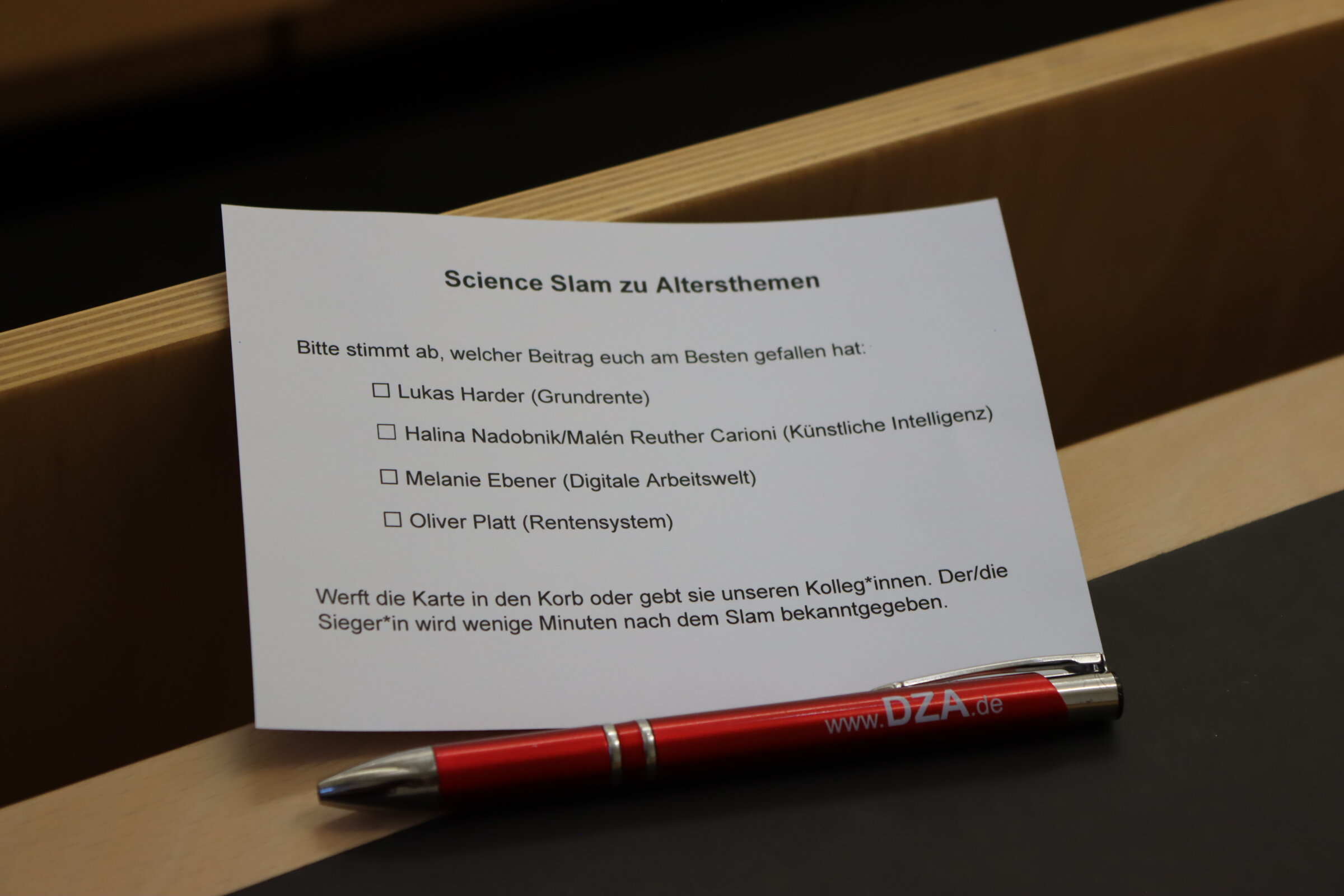 Zu sehen ist die Abstimmungskarte des Science Slam, die zusammen mit einem DZA-Kugelschreiber auf einem Platz im H&ouml;rsaal liegt.